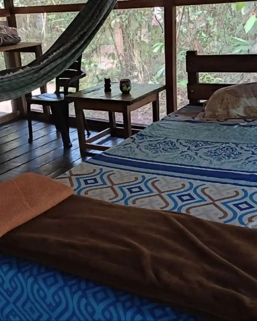 Inside an individual bungalow at the Amazonian solo retreat in Peru — wooden bed with patterned blanket, hammock, desk and open-sided jungle view in Loreto