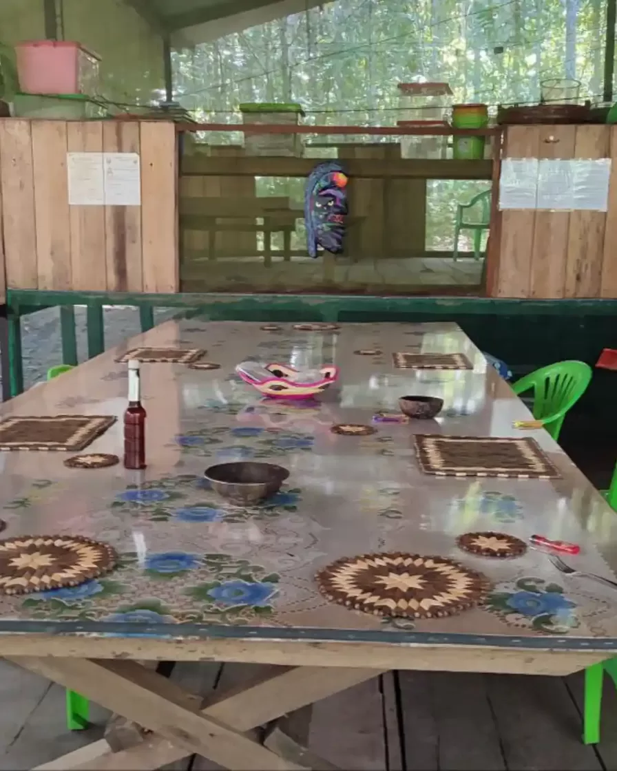Interior of the retreat centre in the Peruvian Amazon — communal table with traditional woven mats, ceremonial mask on the wall and jungle visible through the screens